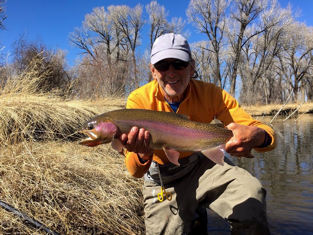 A man with a large trout he caught in the river behind him.