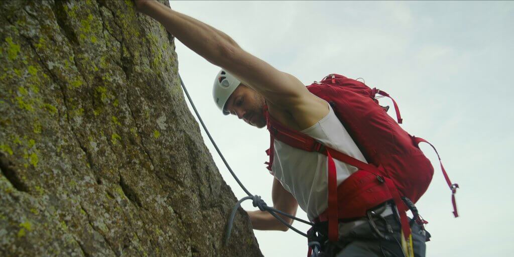 A rock climber wearing a red backpack reaches his hand above as he climbs Devils Tower. 